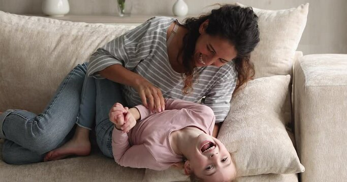 Happy young hispanic mother tickling little laughing kid daughter, resting together on comfortable couch. Joyful sincere two female generations family having fun, enjoying carefree leisure at home.