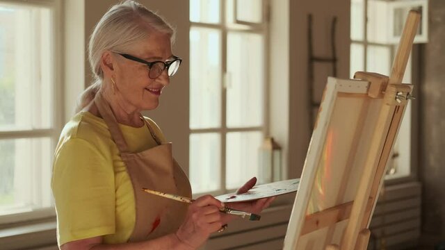 Senior Artist, Art Workshop, Sun Rays, Painting Woman. Happy senior woman artist paints a picture on canvas while standing in a sunny workshop