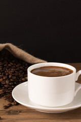 Coffee cup and saucer on wooden background with roasted coffee beans in a burlap sack.