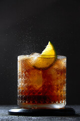Cuba Libre or Long Island Cocktail with rum, cola and lime in glass. Soda spray on dark background. Refreshing summer drink
