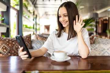 Pretty young woman sitting in cafe at table and use smartphone for video call conversation on table