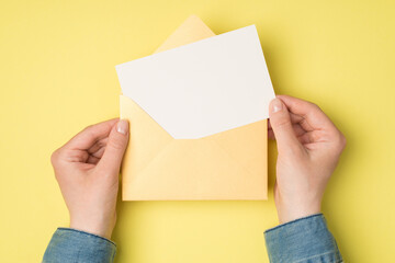 First person top view photo of female hands holding open yellow envelope with white paper card on isolated yellow background with blank space