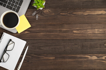 Top view photo of cup of drink yellow sticker note on laptop plant white pen clips and glasses on notepad on isolated dark wooden table background with copyspace