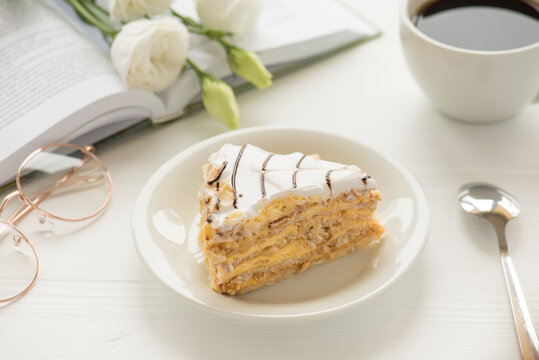Photo of piece of cake on saucer dessert spoon glasses white cup of coffee and flowers on open book on white table