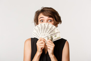 Close-up of excited happy girl holding money near face and peeking at camera thrilled, standing...