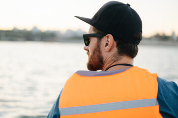 Bearded man in a life vest sitting in front of a large body of water.