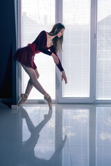 Fototapeta na wymiar Beautiful young ballerina stands and poses in crimson short dress near big window at ballet studio while natural daylight illuminates interior with blue soft ambient light