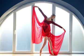 Beautiful young focused ballerina in elegant long red ballet dress in butterfly pose stands in front of big round shaped window at ballet studio  - obrazy, fototapety, plakaty