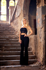 Beautiful young long haired blond woman with serious face expression wearing black top and black long pants stand on staircase inside of old rustic house illuminated with ambient light