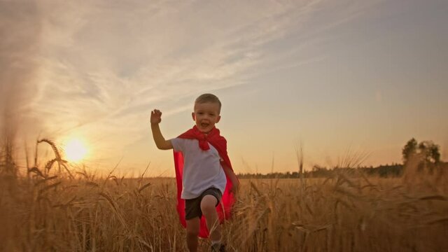 Happy smiling blond boy in red hero cloak runs through meadow wheat field in rays of sunbeams and setting sun. Fun carefree childhood, freedom and dream of flight, children games. Slow motion video.
