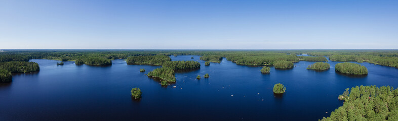 Aerial panorama of Scandinavian green pine tree forest, dark blue lake with small islands. Sunny day, blue sky. Drone photo.
