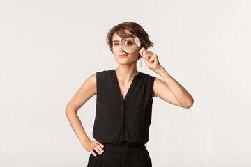 Intrigued stylish woman looking through magnifying glass, smiling thoughtful, standing over white...