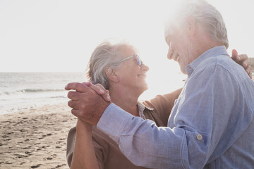 Couple of two old and mature happy seniors enjoying summer dancing together at the beach on the sand with the sunset at the background. Retired and leisure lifestyle.