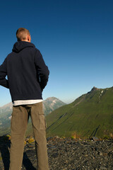 A man from the back, facing green mountains. A hiker contemplates the pastures and snow-capped peaks in summer at dawn.