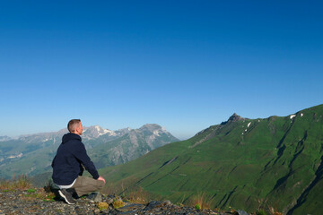 Naklejka premium A man crouching on top of a mountain. Hiker facing a valley of the Alps in summer.