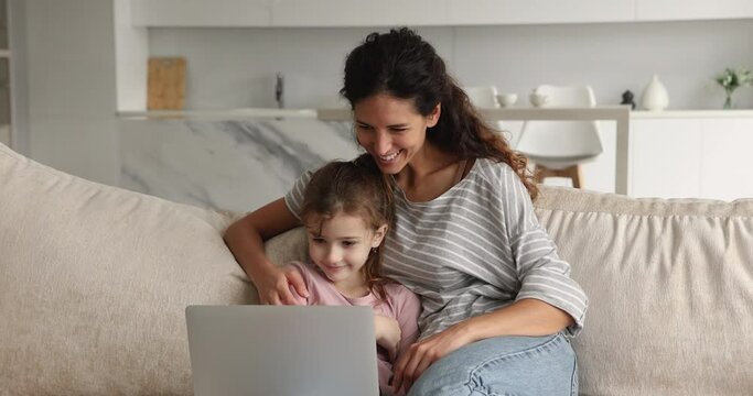 Affectionate caring young mother or nanny watching funny cartoons online on computer with little preteen kid daughter, resting together on comfortable couch at home, leisure weekend activity pastime.