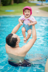 Asian cute baby girl in swimsuit and a pink cap rising up by her father enjoying in the swimming pool on sunny day