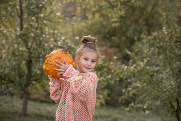 Happy smilling child girl in a knitted sweater is holding in hands ugly orange pumpkin. Deformed...