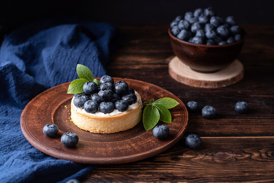 Blueberry tart on a clay plate with tectile on a dark wooden background, summer dessert.