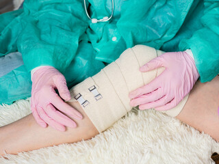 A doctor wearing medical gloves wraps an elastic bandage around the patient's leg and knee. The traumatologist applies an elastic material to the patient