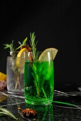 Fototapeta premium Refreshing carbonated drink of bright green color, tarragon and a glass of tonic in the background. Tonic Cocktails. Vertical on a black background