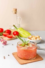 Cold gazpacho soup in a small jar,served with basil and croutons. vegetables for cooking and olive oil in the background. Vertical, on a light background, with space