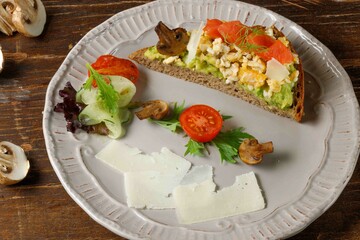 Rye bread toast with guacamole, fried eggs, smoked salmon, and baked champignons. Served on a plate with grilled vegetables and slices of parmesan