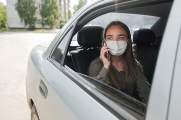 Naklejka premium A beautiful young girl in a mask is sitting in the car and talking on the phone. Protective mask against coronavirus.