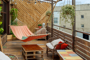 Hammock hangs on wooden terrace with table, flower pots and chairs. Old weathered buildings in the background. Concept of paradise corner in industrial district of big city. - obrazy, fototapety, plakaty