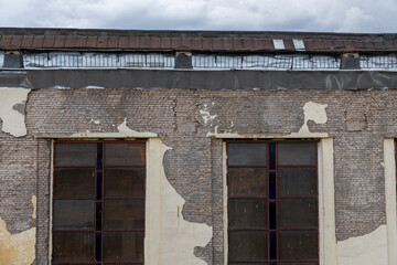 Close-up view of old weathered industrial building with grey brick wall, rusty roof and remains of paint. Grey cloudy sky above the top of building. Old city architecture theme.