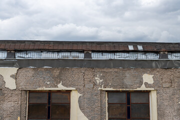 Close-up view of old weathered industrial building with grey brick wall, rusty roof and remains of paint. Grey cloudy sky above the top of building. Old city architecture theme.
