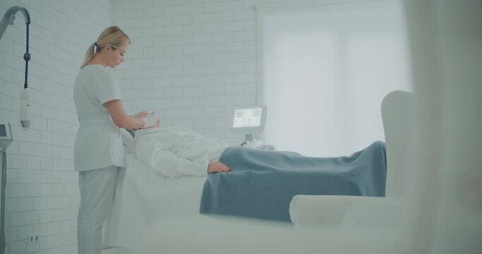 A female client in a bathrobe is laying in a cosmetology bed in a clinic while a female cosmetologist is using a cosmetic pencil to paint white stripes on her cheek.