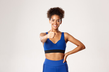 Fototapeta na wymiar Healthy and fit african-american sportswoman, showing thumbs-up in approval, recommend gym or workout app, standing against white background