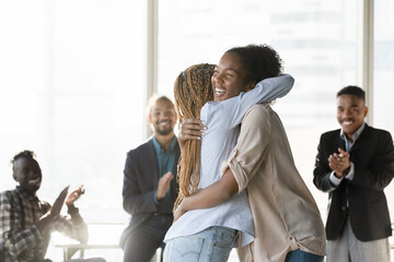 Fototapeta na wymiar Happy young african ethnicity woman cuddling colleague, congratulating with promotion or welcoming newbie at workplace, joyful diverse teammates greeting employee with successful career in office room