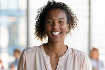 Joyful beautiful young african american female worker manager employee looking at camera, posing in modern office room or holding distant video call conversation with client, successful career concept