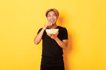 Carefree happy asian guy with blond hair, watching comedy, laughing and eating popcorn, standing...
