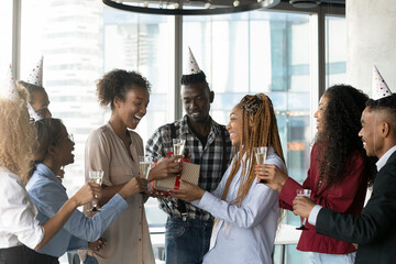 Joyful mixed race african workers in paper party hats congratulating sincere laughing biracial colleague with happy birthday, presenting wrapped gift box, wishing all the best in modern workplace.