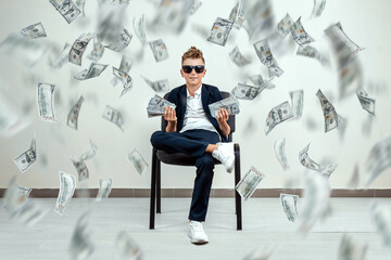 A rich boy with glasses sits on a chair and holds a bundle of dollars in his hands. Knowing how to make money. Money rain, Financial education, young millionaire, investments, loans, winnings.