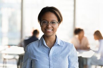 Head shot portrait of smiling millennial african ethnicity businesswoman in eyeglasses posing in modern office room. Confident skilled young biracial employee manager worker looking at camera.