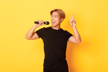 Carefree handsome asian guy performing song, singing into microphone and showing peace gesture, standing yellow background