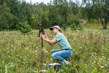 Young woman scientist biologist zoologist sets camera trap for observing wild animals in summer taiga forest to collect scientific data Environmental protection, monitoring of rare and endangered anim - obrazy, fototapety, plakaty