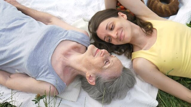 Beautiful old grandmother with grey hair and face with wrinkles is lying on white carpet with her adult daughter on green grass during picnic outdoors, mother's day, happy retirement