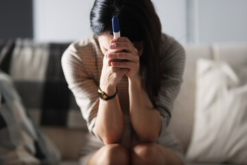 Upset woman is sitting on couch and holding pregnancy test