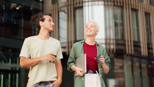 Medium shot of young couple, two friends or colleagues with takeaway coffee and laptop walking down street, talking and discussing city after work. Together again concept