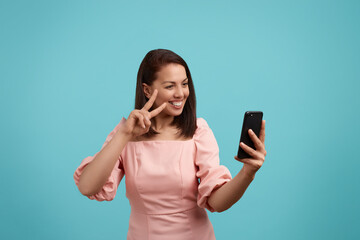 Happy smiling european woman with dark hair in pink dress takes selfie, shows v sign or peace gesture at phone camera, sends good positive vibes, enjoys online video call, isolated on blue background.