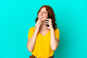 Teenager reddish woman using mobile phone isolated on blue background looking up while smiling