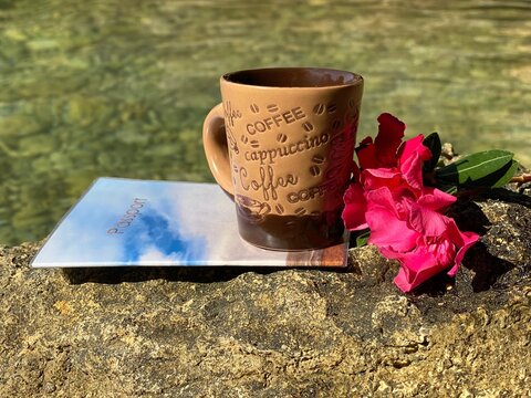 Cup of coffee with pink  flowers on rock face against transparent sea water. Happy summer vacation. Passport for travel. Journey, adventure, summer, tourism. Relaxing holiday on sea shore. 