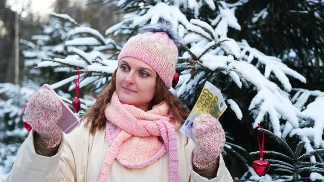 A woman with a phone and money in euros at a Christmas tree in a winter snow-covered forest