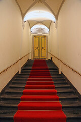 Black stone staircase, with red carpet in the center, leading to a closed decoreted door in an ancient building. Rolli palace, Genoa, Italy