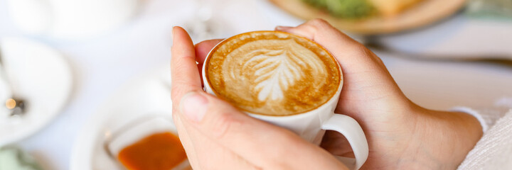 female hands holding a cup of fresh brewed coffee with beautiful leaf latte art from foam on the background of the table with a plate of food. the concept of morning breakfast and brunch. banner